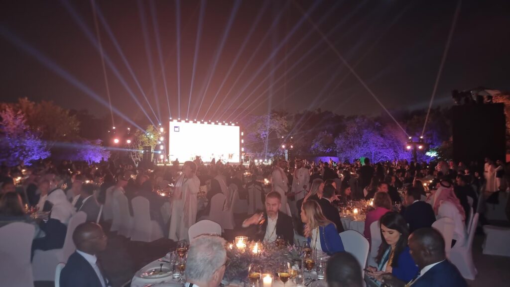 Outdoor gala event at night with large audience, stage screen, and light beams illuminating the venue