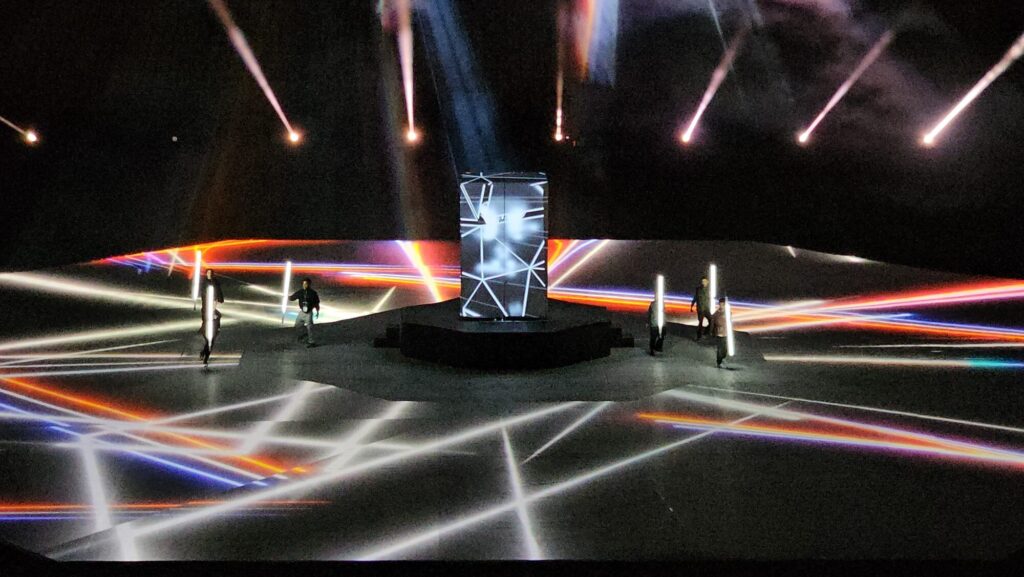 Stadium stage with dynamic light streak projections, central structure, and performers holding illuminated rods