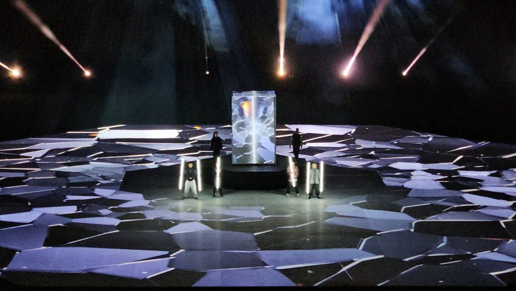 Stadium stage with fragmented floor visuals, central illuminated structure, and performers holding light elements