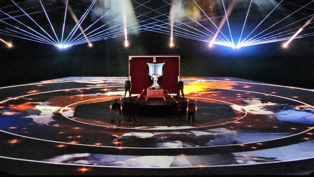 Central stage with illuminated trophy, circular floor projections, and laser beams during a live stadium ceremony