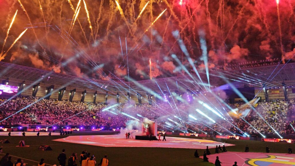 Stadium filled with audience during a live ceremony featuring fireworks, laser lights, and central stage performance