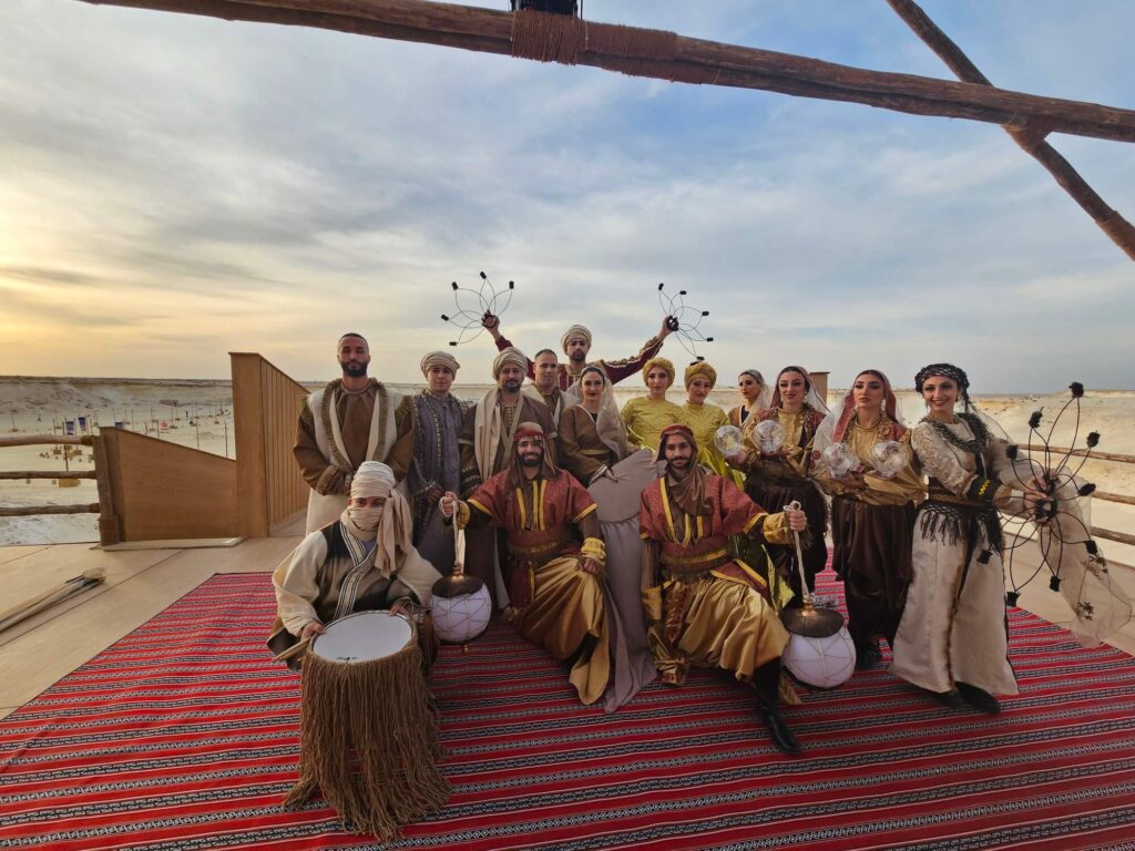 Group of performers in traditional attire posing together on a patterned carpet in a desert setting, some holding musical instruments and props