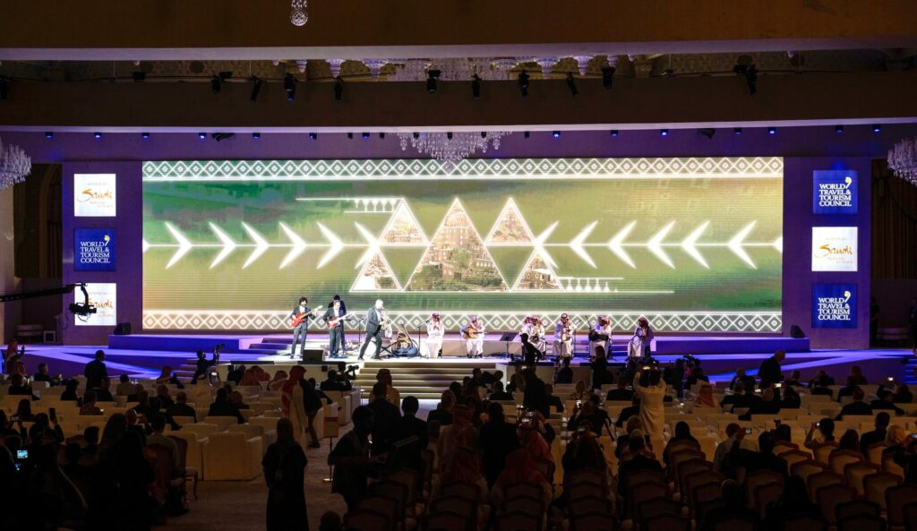 Large-scale live event with band and traditional Saudi musicians performing on stage in front of a seated audience