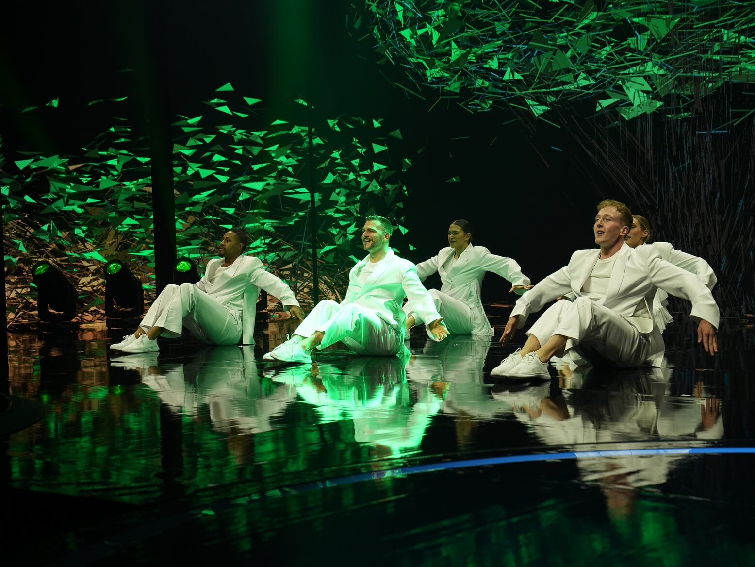 Contemporary dancers in white seated on a reflective stage under green lighting with abstract digital visuals in the background