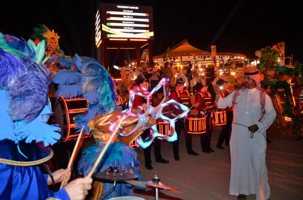 Nighttime street performance with LED drummers, performers in colorful feather costumes, and a man in traditional attire engaging with the scene