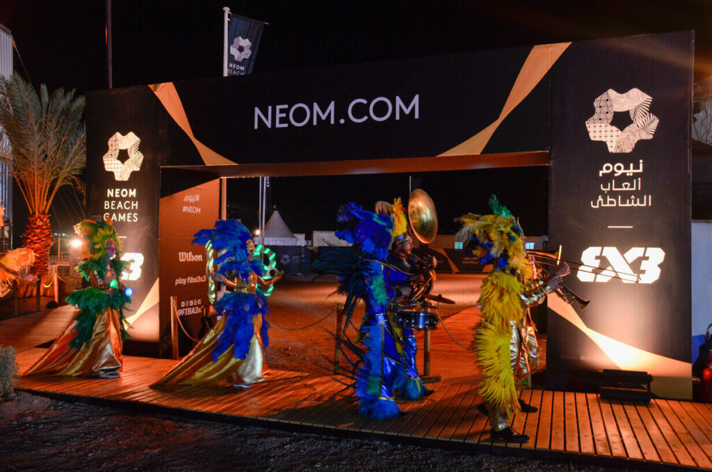 Colorful parade performers with feathers and instruments passing under a NEOM Beach Games entrance arch at night