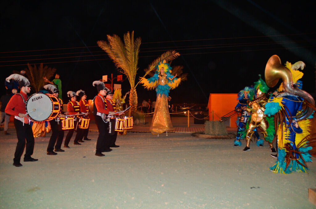 Nighttime performance with LED drummers facing colorful feathered dancers and a stilt performer under palm trees